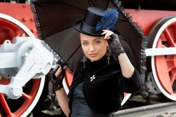 Fototapeta na wymiar Beautiful girl in a black dress and hat near an old steam locomotive and big iron wheels. Blond beauty. Vintage portrait of the last century, retro journey.