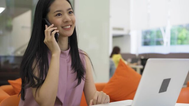 Cheerful Asian businesswoman on phone call alone in modern office buildings. Happy smiling business startup female entrepreneur using laptop - 4K High Quality Footage