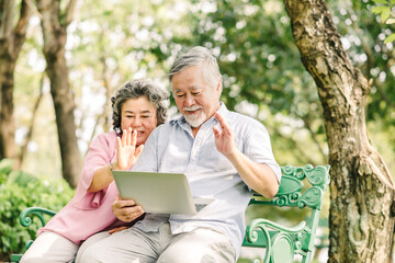Happy senior asian couple waving hand to greet the love one while using laptop outdoor in the park