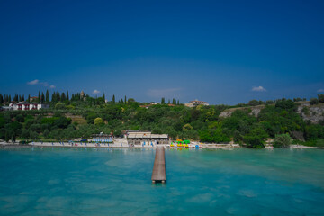 Fototapeta na wymiar Island against the blue sky. Wooden pier, turquoise water. Panoramic aerial view of Lido delle Bionde beach, Sirmione, Lake Garda, Italy.