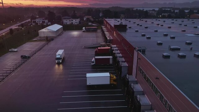Aerial Moving Footage with Multiple Loading Stations with Lorry Trucks at an Industrial Warehouse of Logistics Building. Evening Twilight Sunset with Light Clouds and Pink Hue.