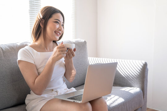 Beautiful young Asian woman working on laptop computer at the living room, Drinking coffee, Work from home.