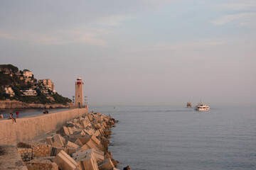 digue avec au fond en perspective un phare en pierre et des bateaux prenant le large au coucher de soleil