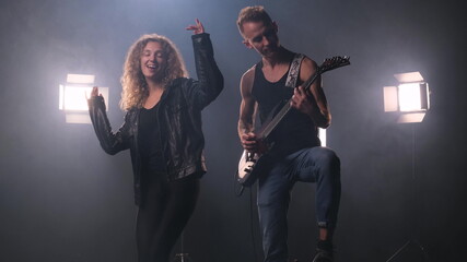 Two rock stars are dancing on stage. The guy in the black T-shirt plays the electric guitar. Woman with lush curly hair in leather jacket dances around him and enjoys music. fog around
