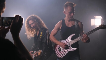 Fan takes phone pictures of two rock stars playing their guitars on stage surrounded by smoke. There is a lot of light. man in black T-shirt and woman in leather jacket play electric guitars