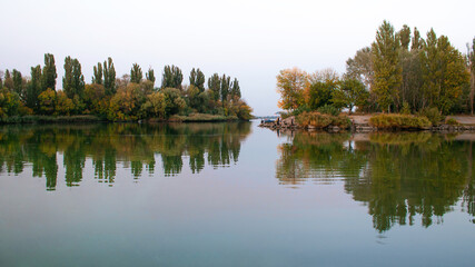 Fototapeta na wymiar Autumn park with a river or lake. Calm and clean water surface. On the shore, people are fishing.