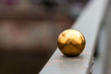 Small gold metal ball on a dark railing. Old matt shabby sphere as a decorative element of the fence. Blurred background, copy space.
