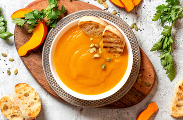 Creamy pumpkin soup in a white bowl, parsley and pumpkin pieces on a gray concrete table top view. Autumnal traditional food. Thanksgiving food.