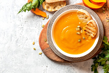 Creamy pumpkin soup in a white bowl, parsley and pumpkin pieces on a gray concrete table top view. Free space for text. Autumnal traditional food. Thanksgiving food. Copy space.