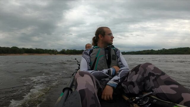 Two men cruising on motorized fishing kayak upstream through Wisconsin river in overcast day while looking for fishing spot.