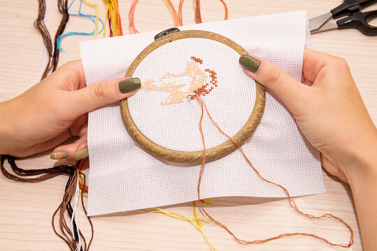 Women's hands hold a white canvas of cross-stitch embroidery in a hoop with a needle, against the background lie the scissors and multicolored floss threads