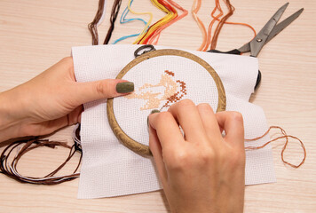 Women's hands hold a white canvas of cross-stitch embroidery in a hoop with a needle, against the background there are multicolored floss and scissors © Наталья Анюхина
