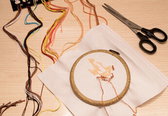 Cross-stitch embroidery in the hoop lies on a wooden background along with multicolored floss and...