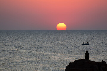The solar disk rises to the horizon. Rock, silhouette, and boat.
