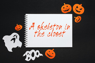 Happy halloween holiday concept. Notepad with text A skeleton in the closet on black background with bats, pumpkins and ghosts