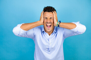 Naklejka premium Young handsome man wearing blue shirt over isolated blue background crazy and scared with hands on head, afraid and surprised of shock