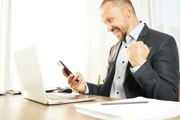 a businessman man in the office has read a news or message in his smartphone and brightly expresses emotions of joy, surprise and happiness with a smile