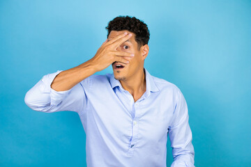 Young handsome man wearing blue shirt over isolated blue background peeking in shock covering face and eyes with hand, looking through fingers with embarrassed expression