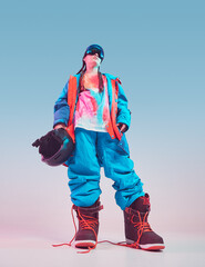 Fototapeta premium Cool sporty young female in stylish colorful winter sportswear with snowboard helmet posing in studio in neon light