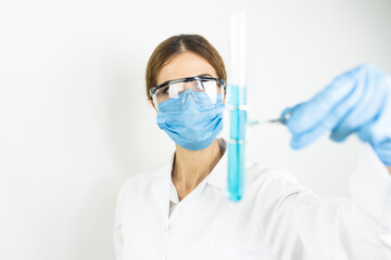 Young attractive concentrated female scientist in protective eyeglasses, mask and gloves dropping a blue liquid substance into the test tube with a pipette in the scientific chemical laboratory   