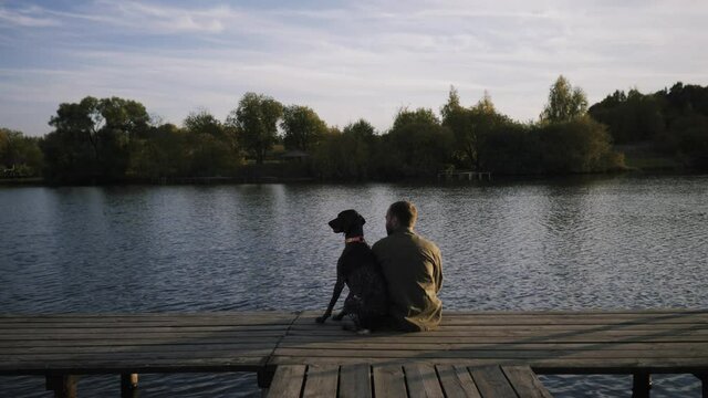 young guy sitting with dog on the pier and admiring the beauty of the lake