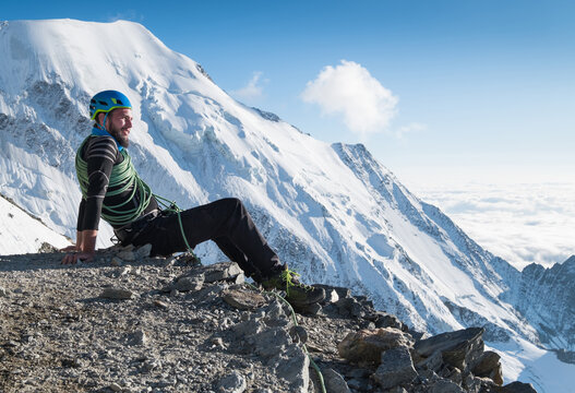 Bearded Climber in a safety harness, helmet, and on body wrapped climbing rope with sitting at 3600m altitude with picturesque Aiguille de Bionnassay mountain during Mont Blanc France route