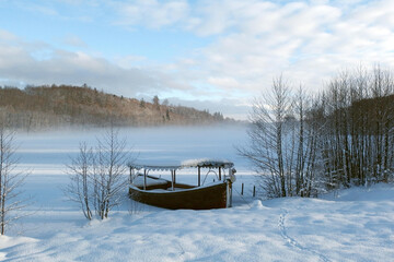 Snowy winter landscape with wooden boat standing on bank of frozen misty lake