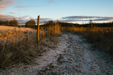 An empty road in the countryside, covered by a thin layer of snow, the morning sun shines through the field behind the road