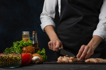 The professional chef in black apron cuts with knife chicken fillet on black chopped board on dark...