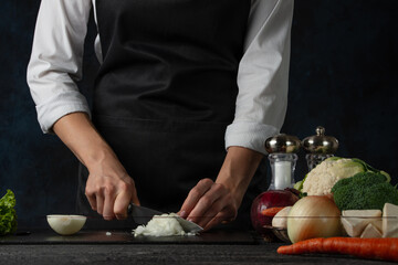 The professional chef in black apron cuts with knife onion on black chopped board on dark blue background. Backstage of preparing restaurant meal for dinner. Food concept. Close-up.