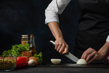 Close-up view of chef in black apron cuts with knife onion on professional black chopped board on dark blue background. Backstage of preparing meal with natural ingredients for cooking. Food concept.