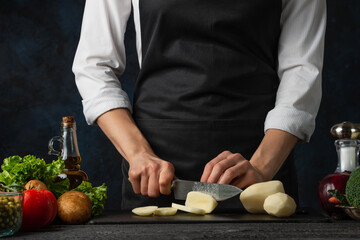 The professional chef in black apron cuts with knife potato on black chopped board on dark blue background. Backstage of preparing restaurant meal for dinner. Food concept. Close-up.