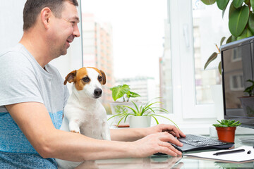 Caucasian man sits at a computer by window with houseplants, smiles, dog sits on his lap, put his paw on owner's hand