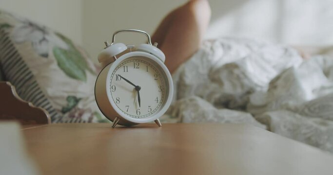 The man is woken up by an alarm clock at six in the morning. The man's hand stops the alarm clock to sleep longer. Sleeping man reluctantly wakes up to an alarm clock early in the morning.