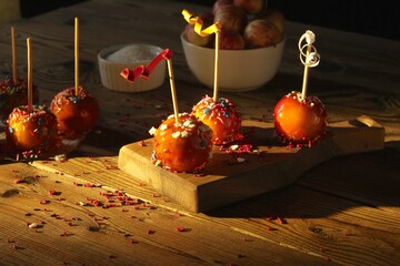 Apples in red caramel, decorated with decor on a wooden board on a dark background. Illuminated by the bright rays of the sun. In the background there are small apples and a plate of sugar