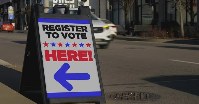 A 'REGISTER TO VOTE HERE' sign on the sidewalk of a large city as traffic passes in the background.	