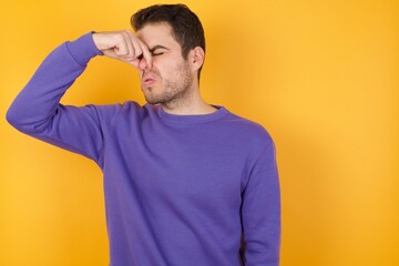 Handsome man with sweatshirt over isolated yellow background smelling something stinky and disgusting, intolerable smell, holding breath with fingers on nose. Bad smell