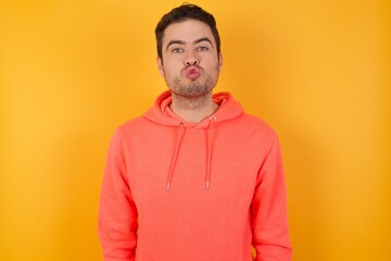 Shot of pleasant looking Handsome man with sweatshirt over isolated yellow background, pouts lips, looks at camera, Human facial expressions