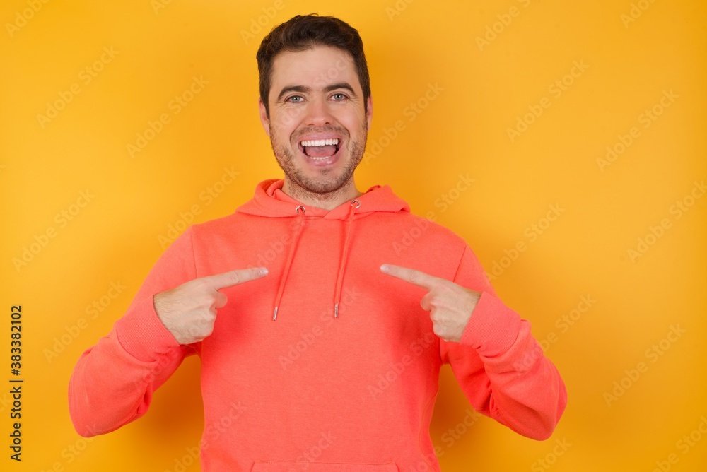 Wall mural Handsome man with sweatshirt over isolated yellow background points at his body, being in good mood after going shopping and making successful purchases - Wall murals