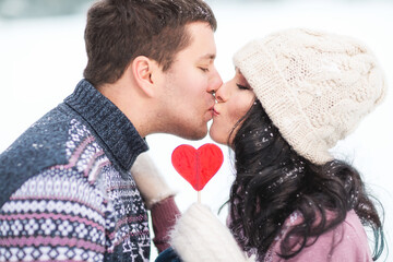 Outdoors lifestyle close up portrait of happy kissing couple in love. Holding in their hands red lollipop in shape of heart. On honeymoon. Wearing knitted hat, mittens and sweaters.  Love concept