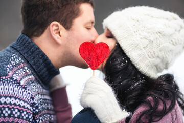 Outdoors lifestyle close up portrait of happy kissing couple in love. Holding in their hands red lollipop in shape of heart. On honeymoon. Wearing knitted hat, mittens and sweaters.  Love concept