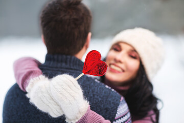 Outdoors romantic portrait of happy hugging couple in love. Girl smiling and holding in her hand red lollipop in shape of heart. Wearing knitted hat, mittens and sweaters. Love concept. Valentine day
