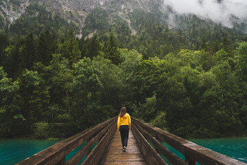 Girl in yellow walking on a bridge over a clear blue river surrounded by green forest