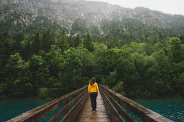 Girl in yellow walking on a bridge over a clear blue river surrounded by green forest