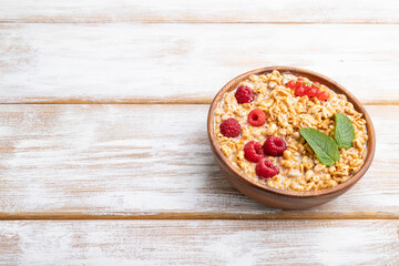 Wheat flakes porridge with milk, raspberry and currant in wooden bowl on white wooden background. Side view, copy space.