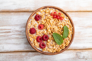 Wheat flakes porridge with milk, raspberry and currant in wooden bowl on white wooden background. Top view, close up.