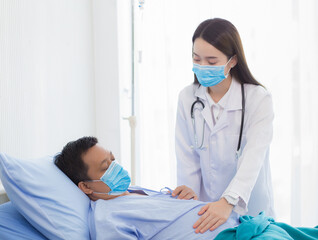 An Asian woman doctor is checking the symptom of a man patient who bed rest in hospital. Both wear a surgical mask to protect Corona virus disease (Covid 19).