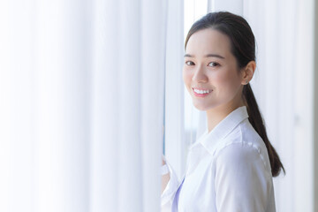 Asian beautiful woman in white shirt is smiling and standing near window with white curtain.