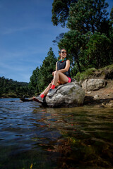 Fototapeta na wymiar Atleta femenino descansando sobre una roca a la orilla de un lago, después de correr en el bosque