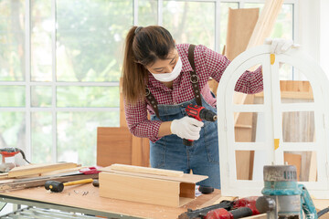 A confident young female carpenter uses a hand drill to assemble the wood window in the carpentry...